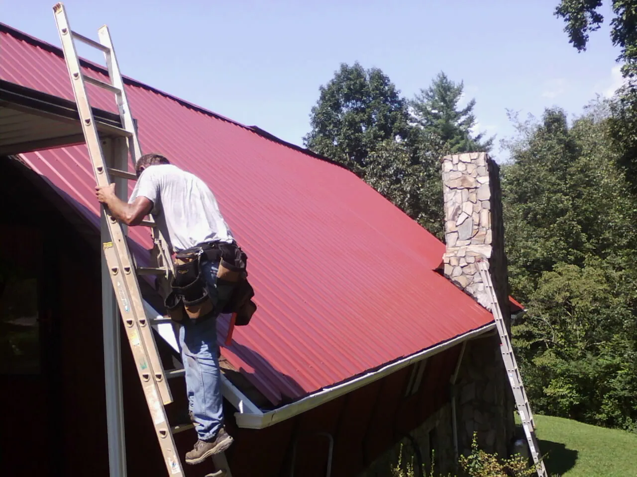 Roofer on a ladder beside a red metal roof and stone chimney — Soffit & Fascia Repair by Roofing Brothers in Huntland